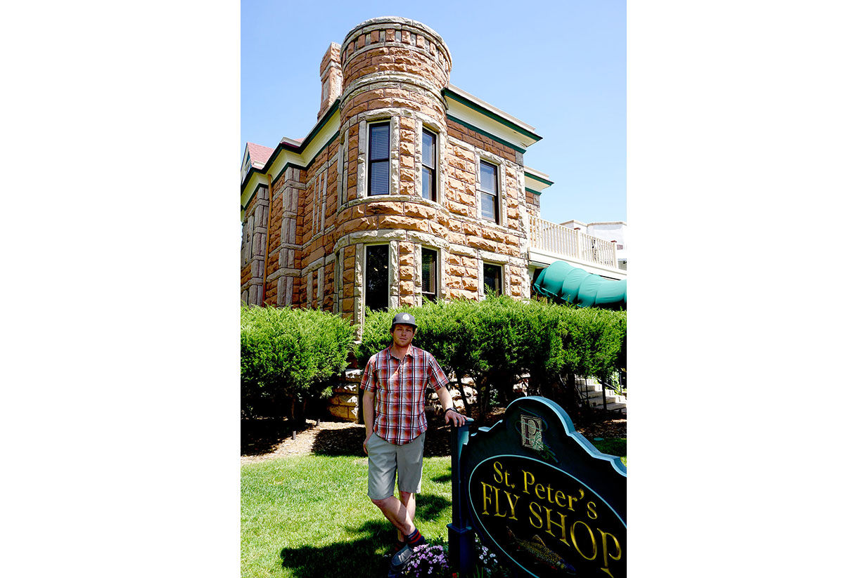 A man stands in front of a brownstone historic building and leans on a sign: St. Peter's Fly Shop